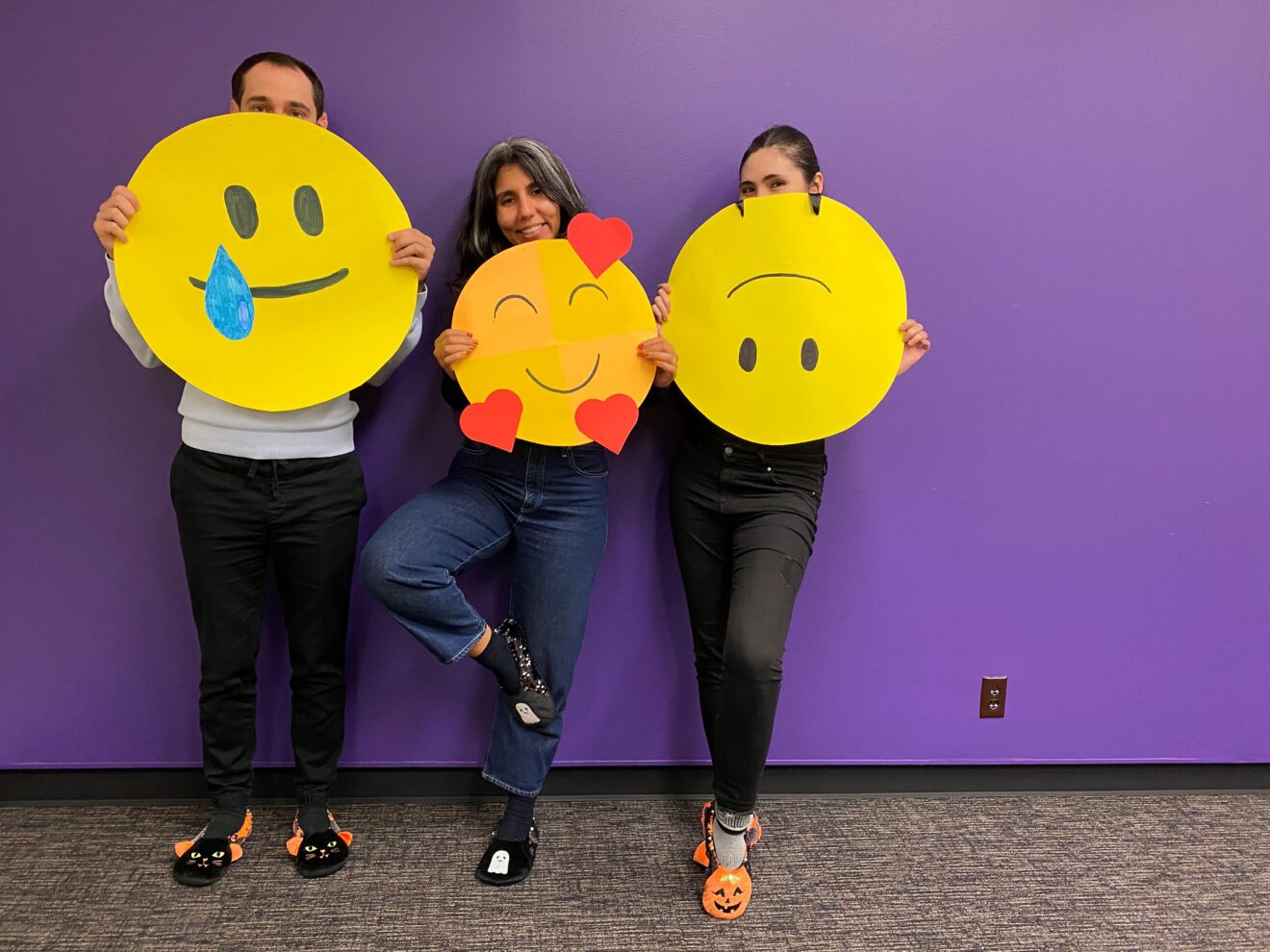Three people lean against a purple wall holding yellow construction paper emojis in front of them to celebrate Halloween.