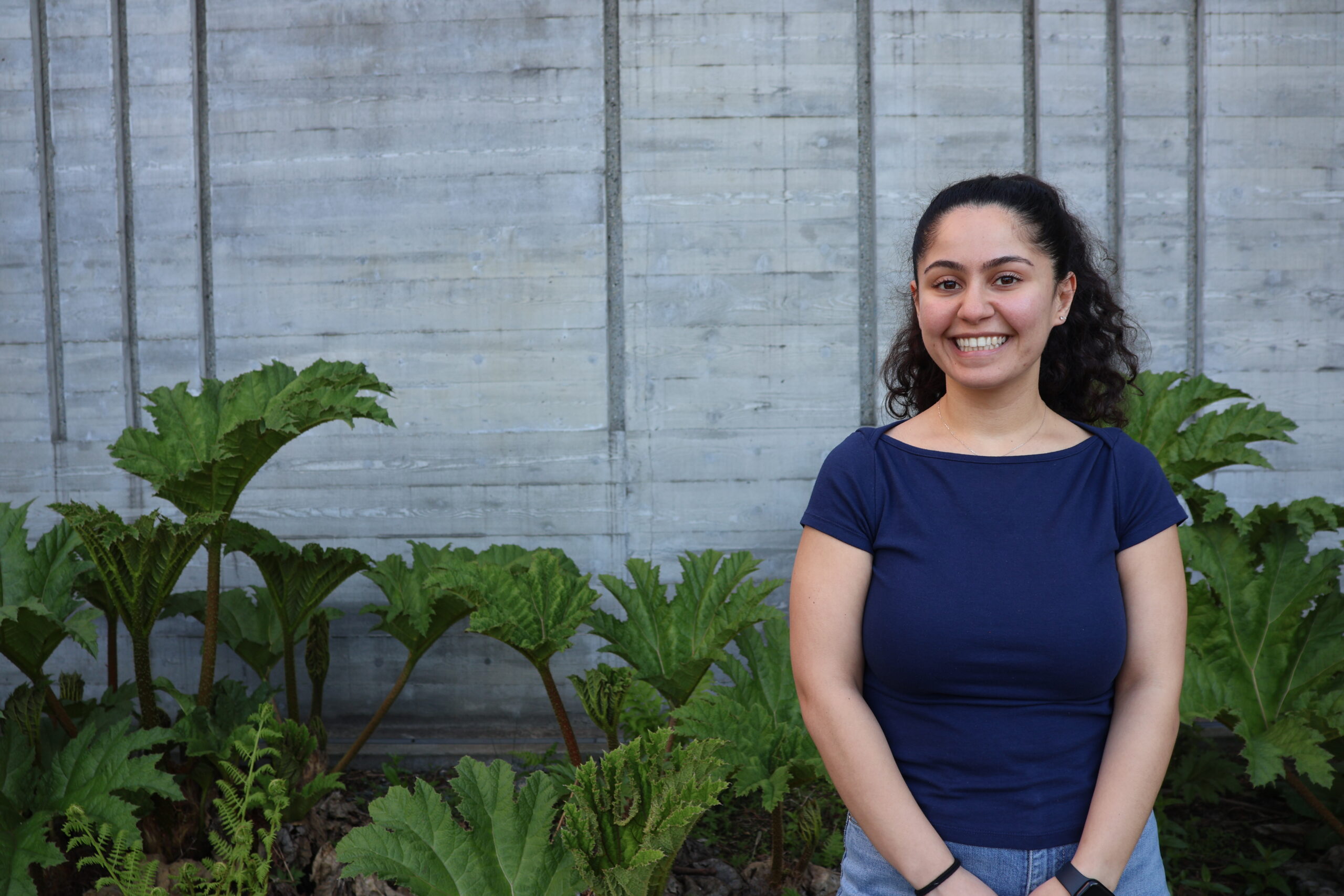 Elissa smiles while standing in front of leafy green plants and a concrete wall.